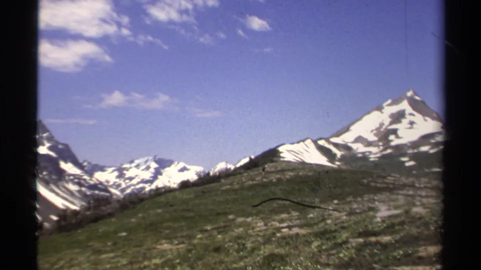 1979 8mm British Columbia mountain view with green alpine ground, snow-covered peaks, and scattered clouds in a blue sky.