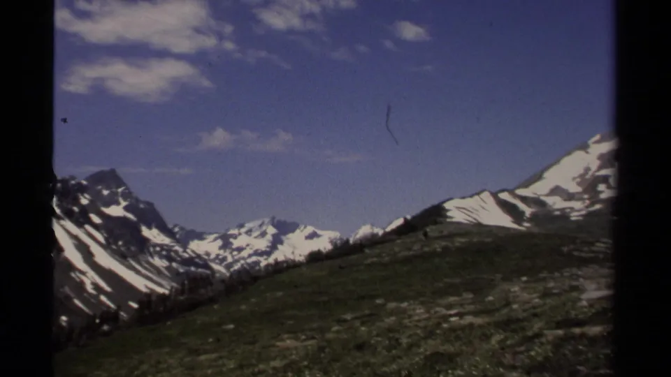 1979 8mm British Columbia landscape showing a grassy slope, dark ridges, and snow-capped mountains under scattered clouds.