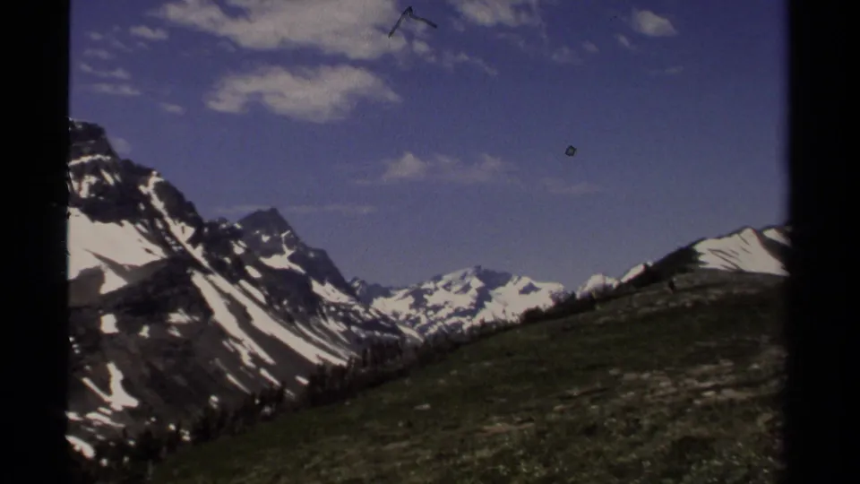 1979 8mm British Columbia view of snow-streaked ridges, distant peaks, and open green mountainside beneath a blue sky.