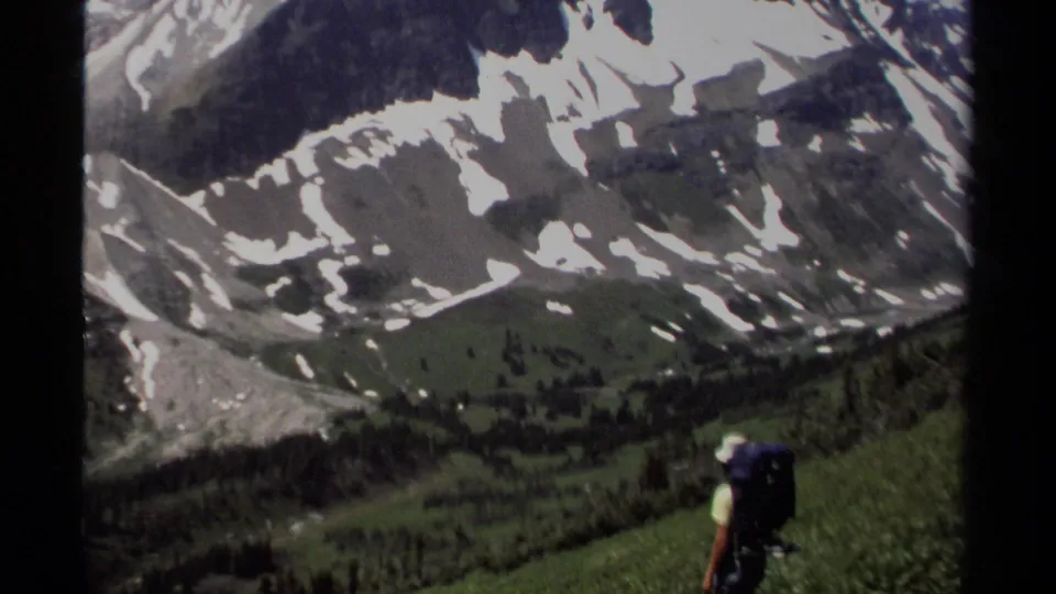 1979 British Columbia 8mm frame of a backpacked hiker on a grassy slope facing snow-dappled mountains.