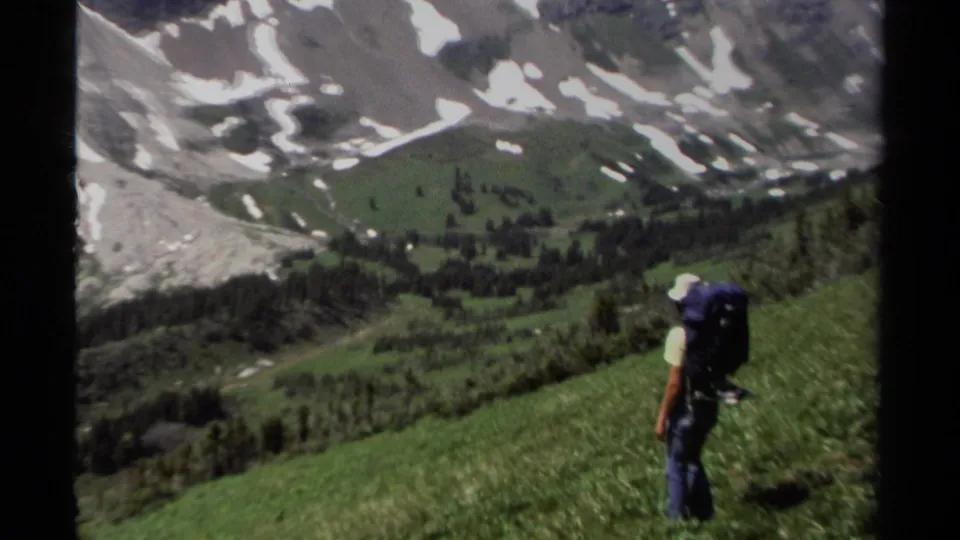 1979 British Columbia frame showing a hiker with a large pack looking across green slopes and snowy mountains.
