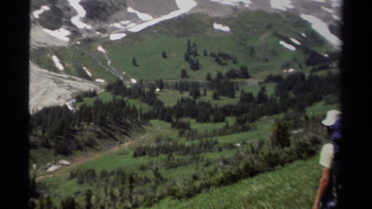 Backpacker on a grassy ridge looking down toward a green valley and dirt road.