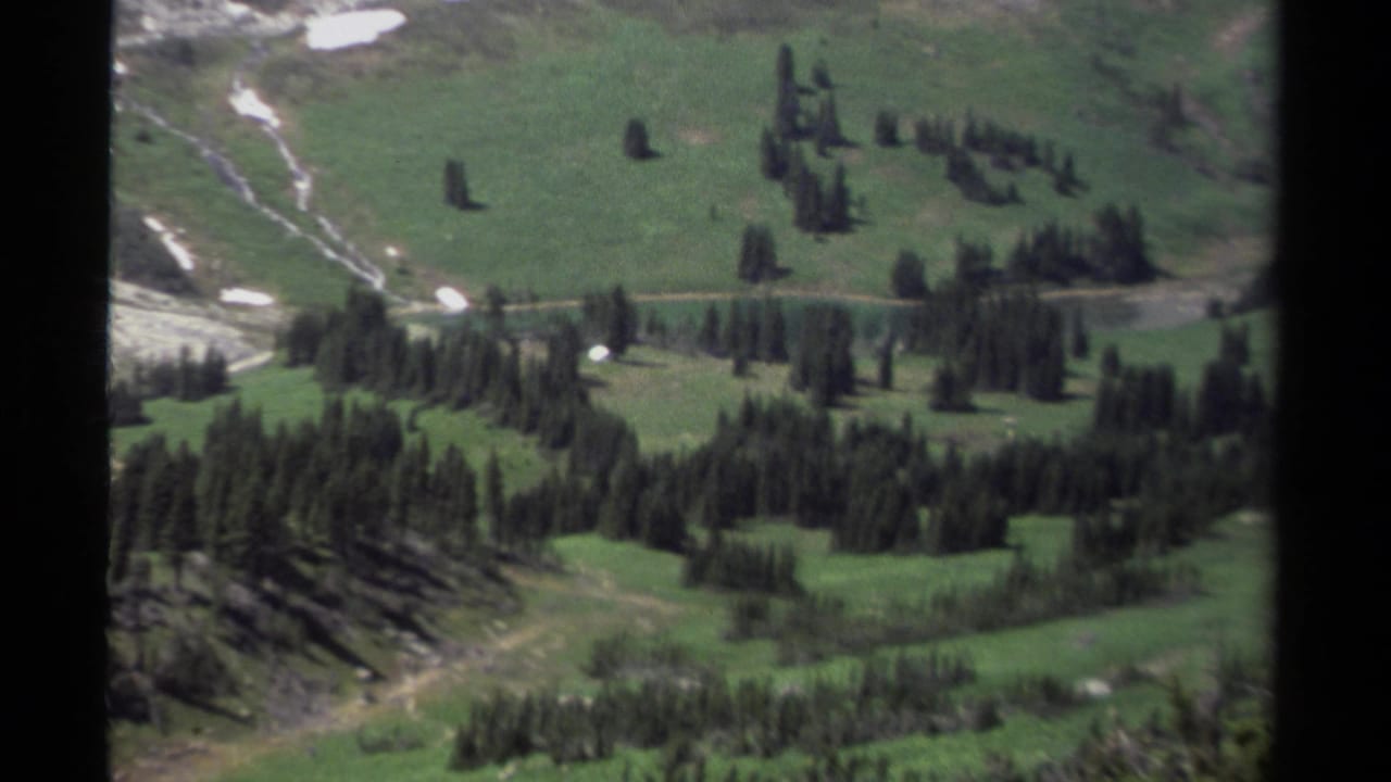 1979 British Columbia 8mm view across a green mountain valley with evergreen stands, a pale dirt road, and snow-dappled terrain.