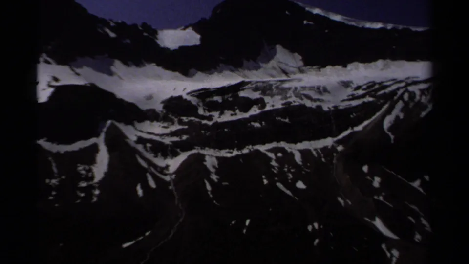 1979 British Columbia 8mm frame showing a darker mountain face, lingering snow, and steep alpine terrain beyond the valley.