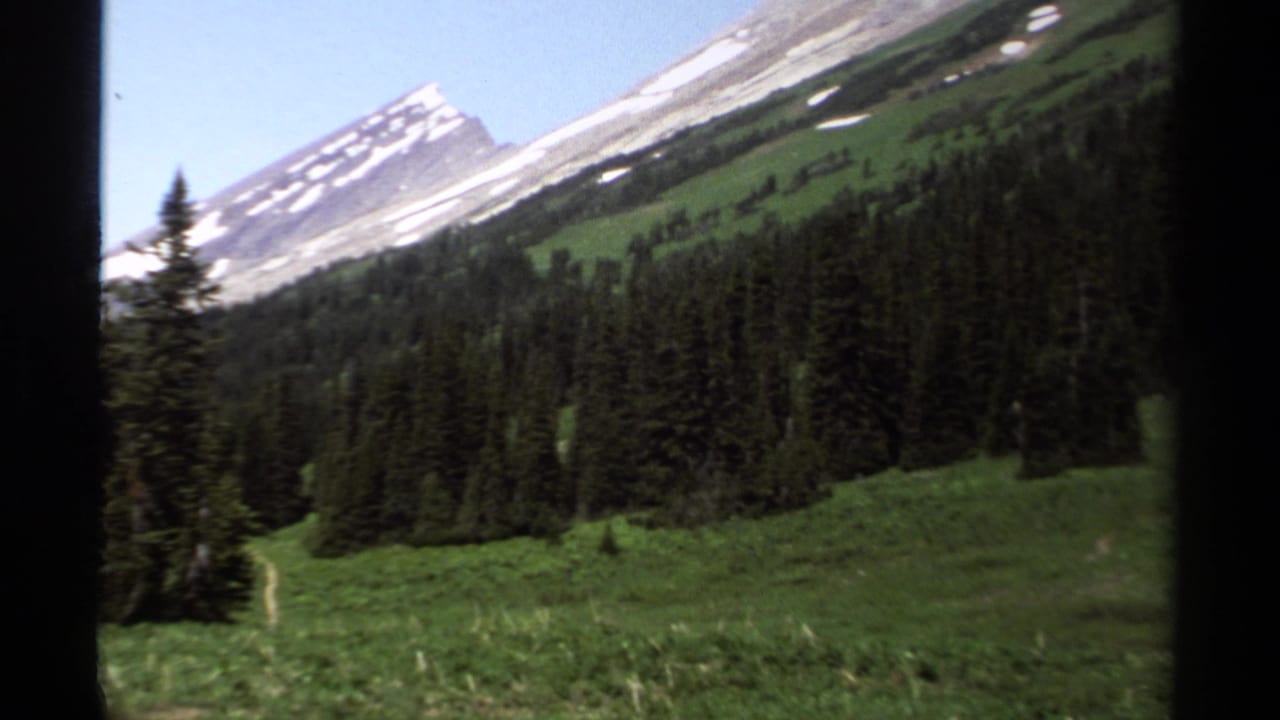 1979 British Columbia landscape frame with conifers, alpine meadow, and a snow-streaked peak partly framed by a vehicle window.