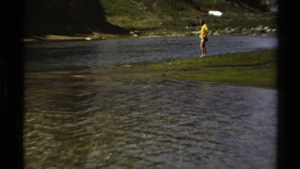 A distant adult man in a yellow shirt stands on a grassy bank across the water in British Columbia 8mm home-movie footage.