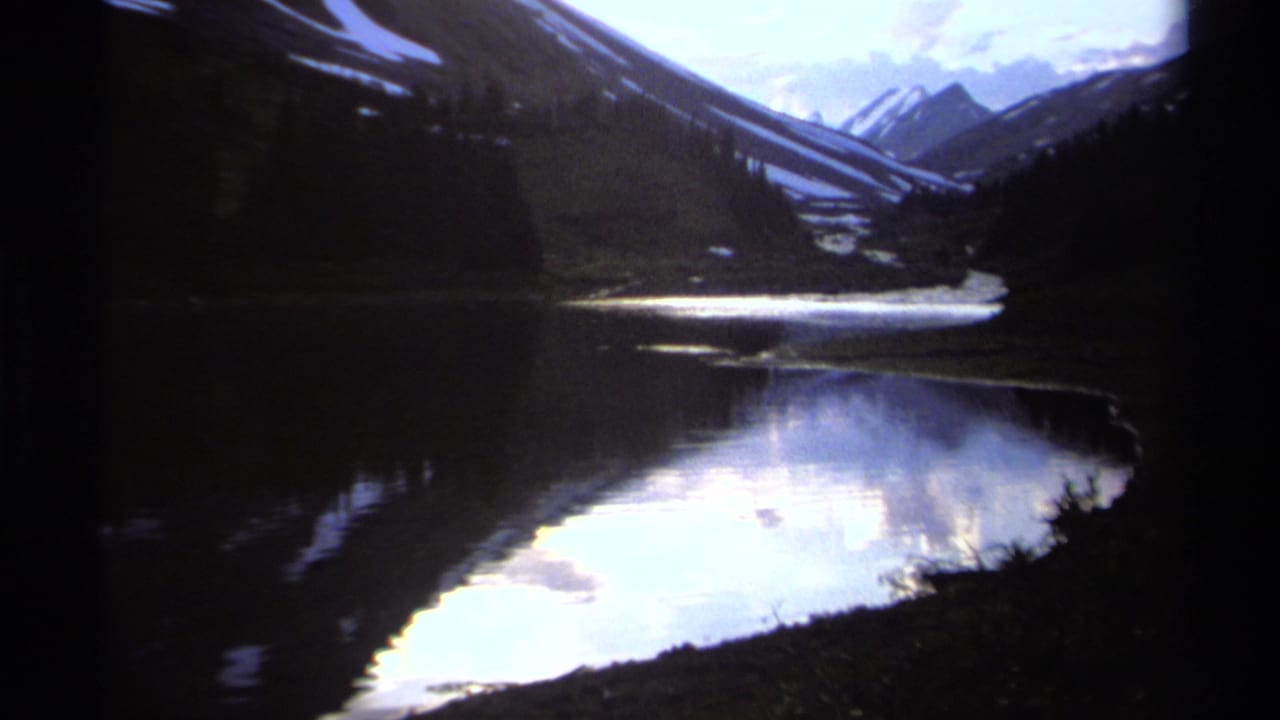 1979 8mm home-movie view of a British Columbia mountain lake with snow-capped peaks reflected in still water.
