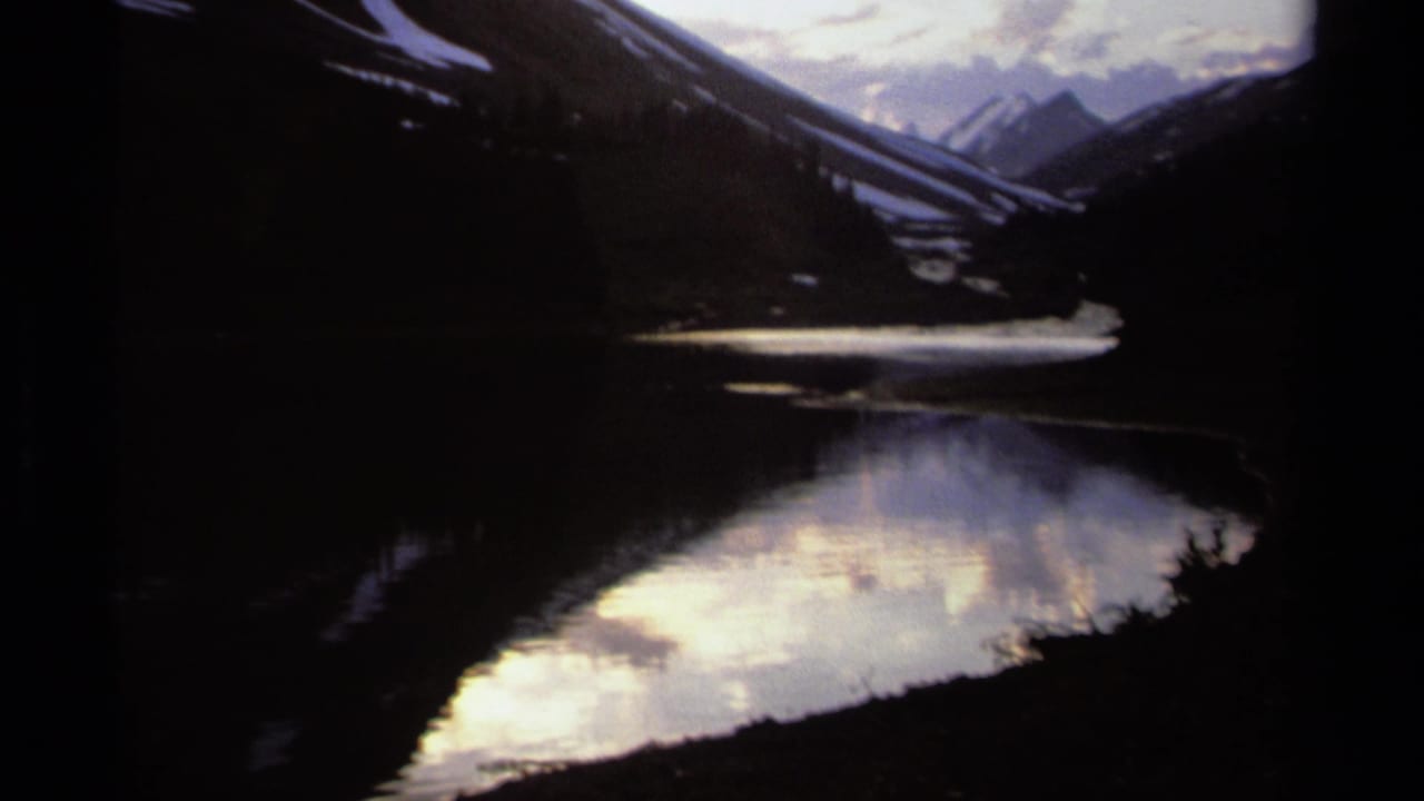 British Columbia mountain lake scene on 8mm film, with dimmer exposure, reflected clouds, and snowy peaks in the distance.