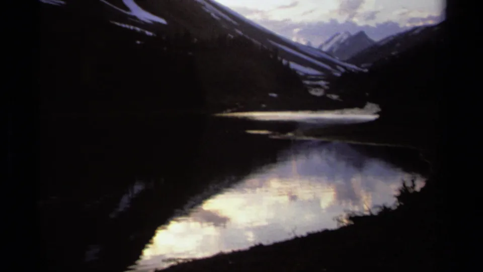 British Columbia mountain lake scene on 8mm film, with dimmer exposure, reflected clouds, and snowy peaks in the distance.