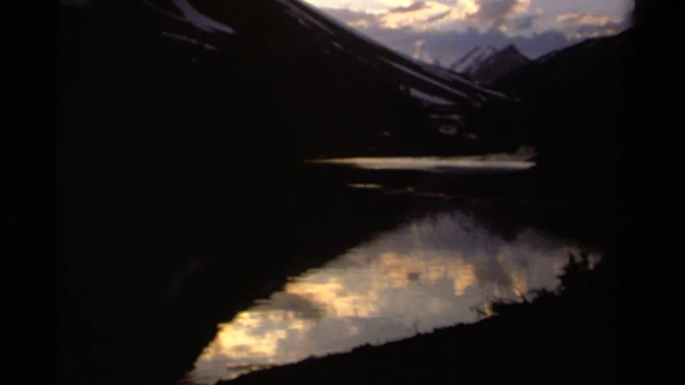 8mm home-movie frame of a British Columbia lake valley with dark mountain slopes and warm cloud reflections on the water.