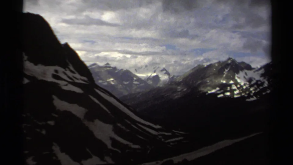 Wide 1979 8mm mountain vista in British Columbia with dark foreground slopes, snowfields and clouded distant peaks.