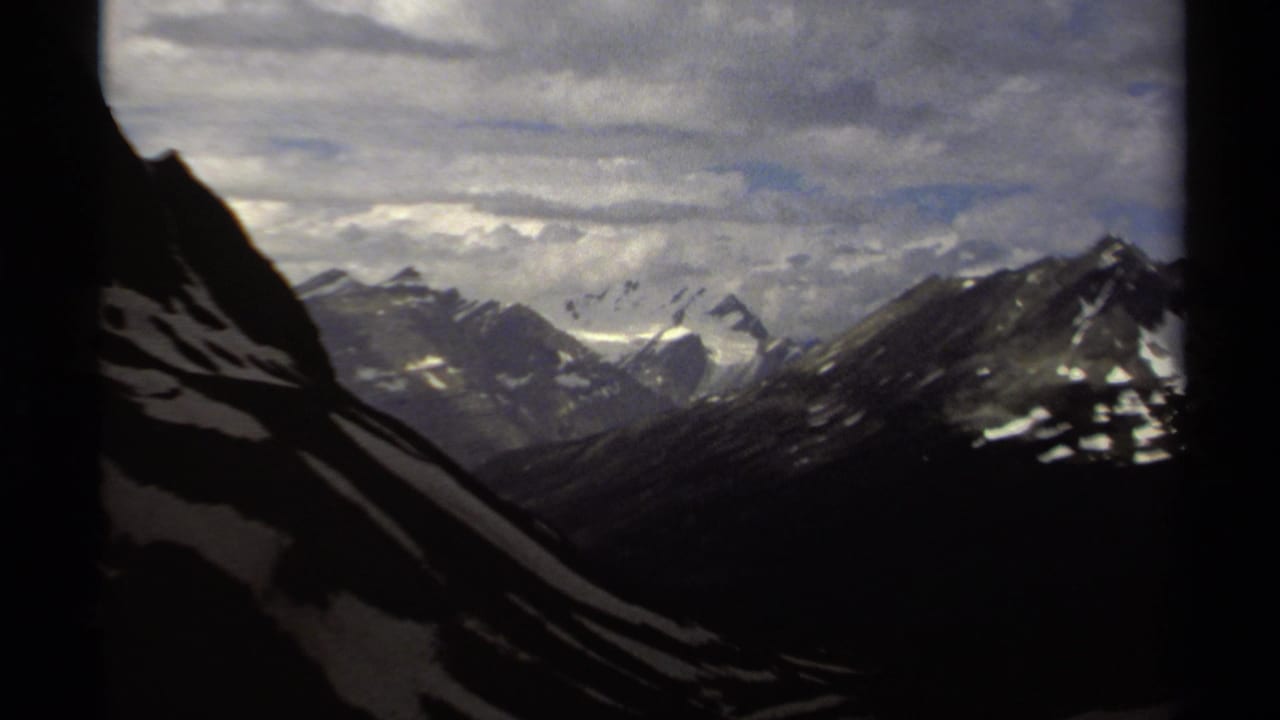 1979 British Columbia 8mm frame showing distant snow-covered mountain peaks under a layered cloudy sky, with dark slopes in the foreground.