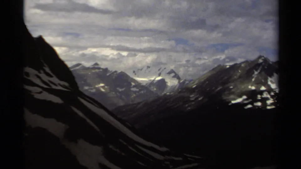 1979 British Columbia 8mm frame showing distant snow-covered mountain peaks under a layered cloudy sky, with dark slopes in the foreground.