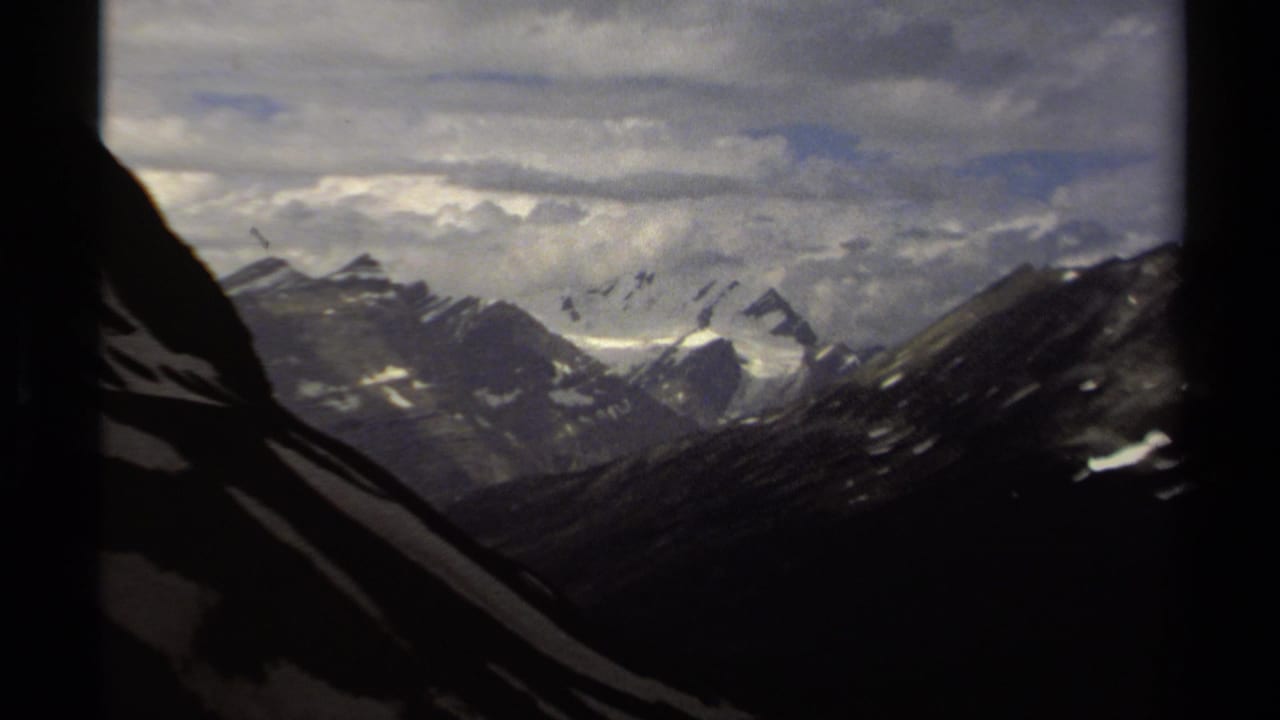 1979 British Columbia mountain view with jagged snow-lined ridges, deep shadowed slopes, and dense cloud layers across the sky.