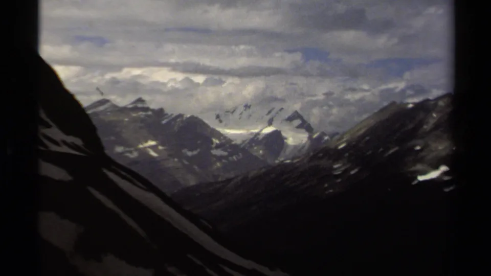 1979 British Columbia mountain view with jagged snow-lined ridges, deep shadowed slopes, and dense cloud layers across the sky.