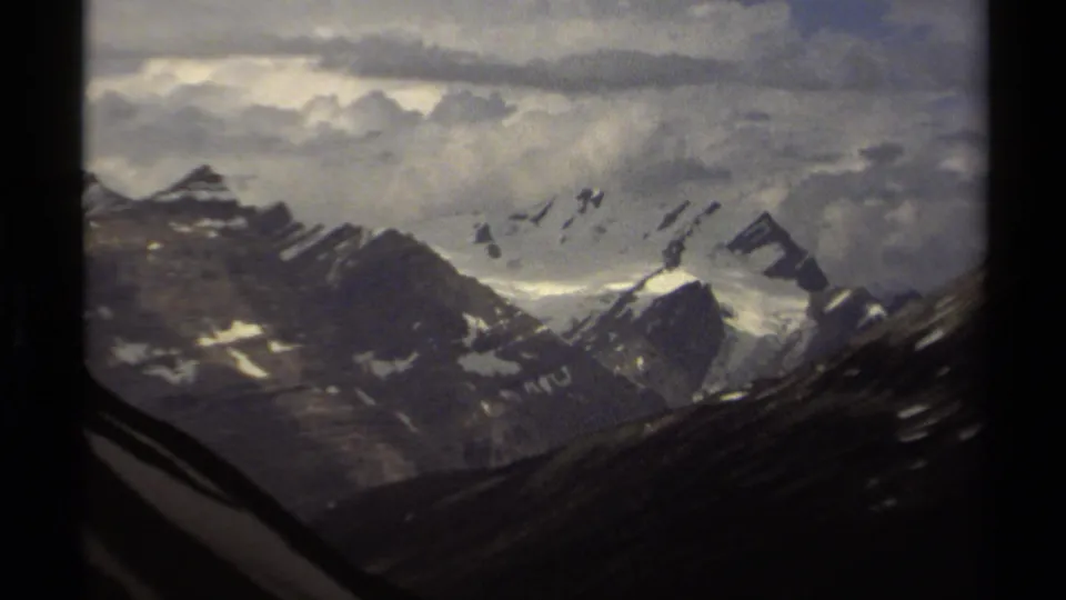 1979 British Columbia frame centered on snow-covered alpine peaks and a bright snowfield beneath textured, layered storm clouds.