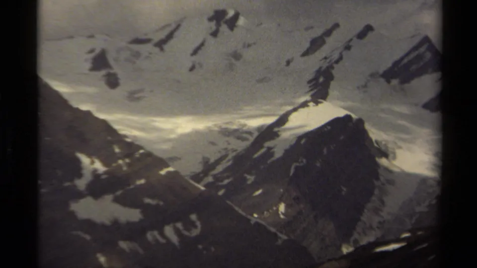 Closer 8mm frame of British Columbia alpine terrain, showing a snow-covered mountain face, dark ridges, and muted archival color.