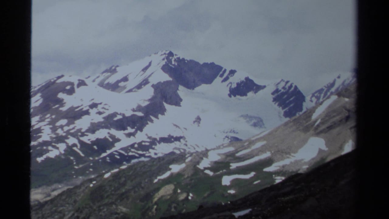 1979 British Columbia mountain view showing snowfields across high peaks, dark ridges, green lower slopes, and a cloudy sky.