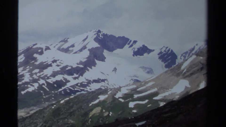 1979 British Columbia mountain view showing snowfields across high peaks, dark ridges, green lower slopes, and a cloudy sky.