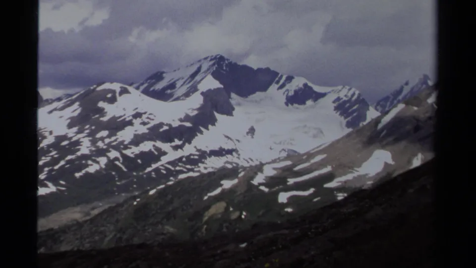 British Columbia 1979 home-movie frame showing a broad snowy mountain range, dark film borders, rocky slopes, and overcast sky.