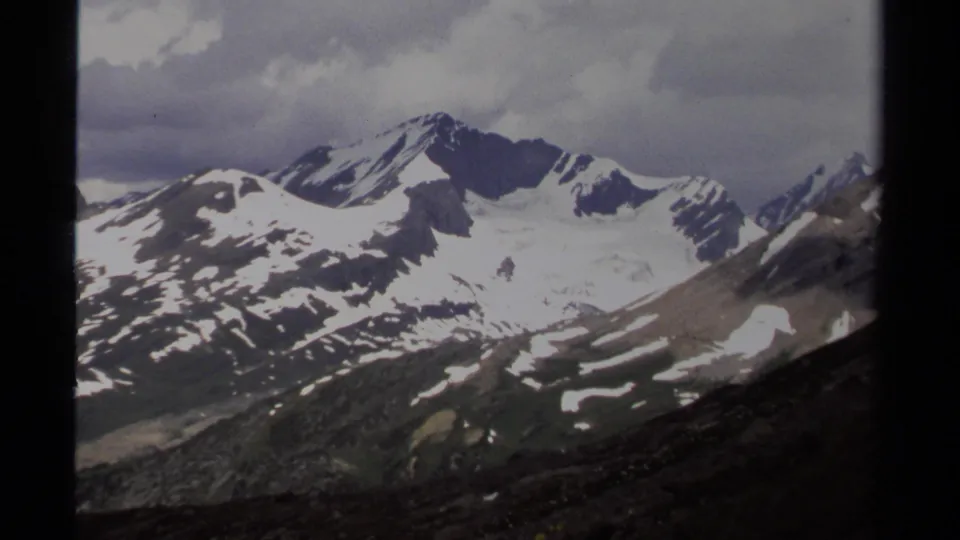 1979 British Columbia 8mm frame with snow-striped peaks, rocky foreground terrain, gray clouds, and soft film grain.