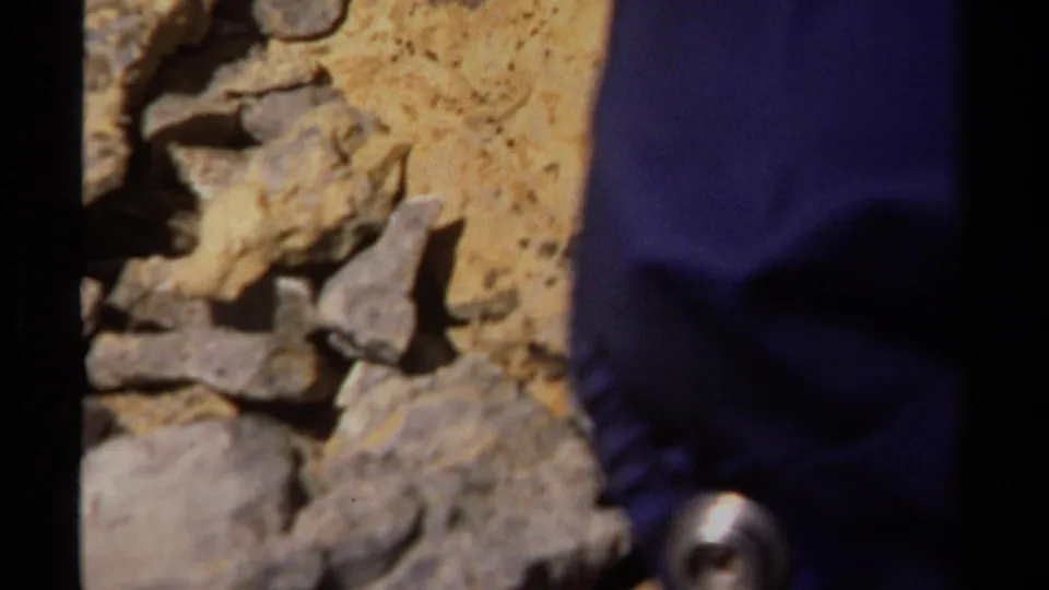 Close 1979 8mm view of rocks and sandy ground with part of a hiker's blue-clad leg entering the right edge of frame.