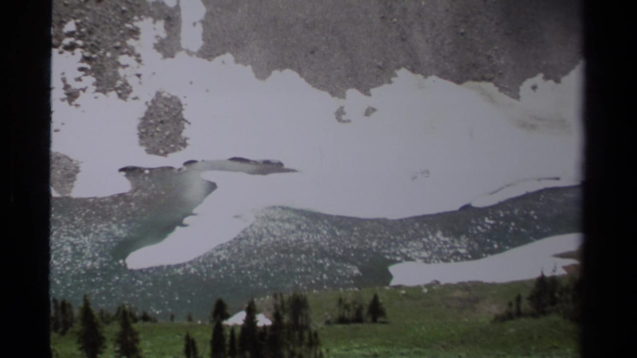 Snow patches on a rocky alpine slope above meadow and evergreens.