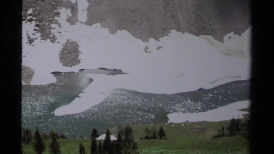 1979 8mm view in British Columbia showing melting snow across a rocky mountain slope above green meadow and scattered evergreens.