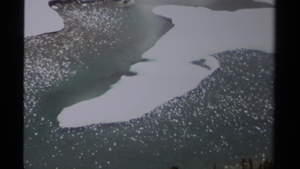 Closer 1979 8mm frame of melting snow bordering turquoise water or creek flow on a high British Columbia mountain slope.