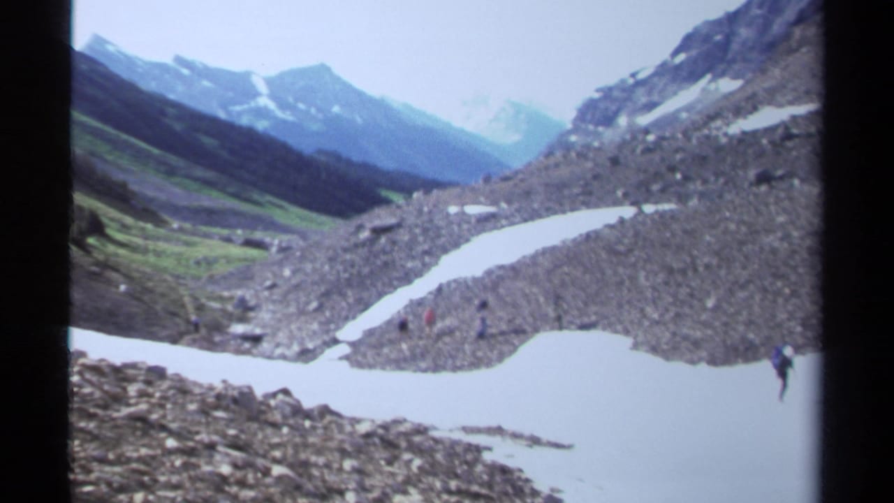 1979 British Columbia 8mm frame showing small hikers in a rocky, snow-dappled alpine valley with layered peaks beyond.