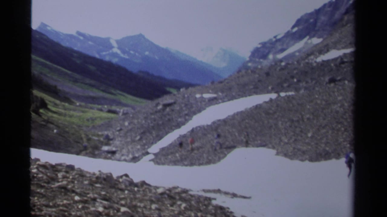 Small figures walk near snow patches in a rocky mountain valley.