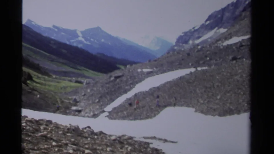 1979 British Columbia 8mm frame of a snow-dappled rocky valley, green mountainside, and distant alpine peaks under a pale sky.
