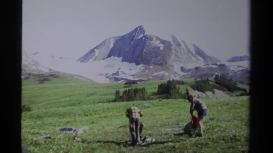 1979 British Columbia frame showing two hikers or researchers working in a grassy alpine meadow below a snow-capped mountain.