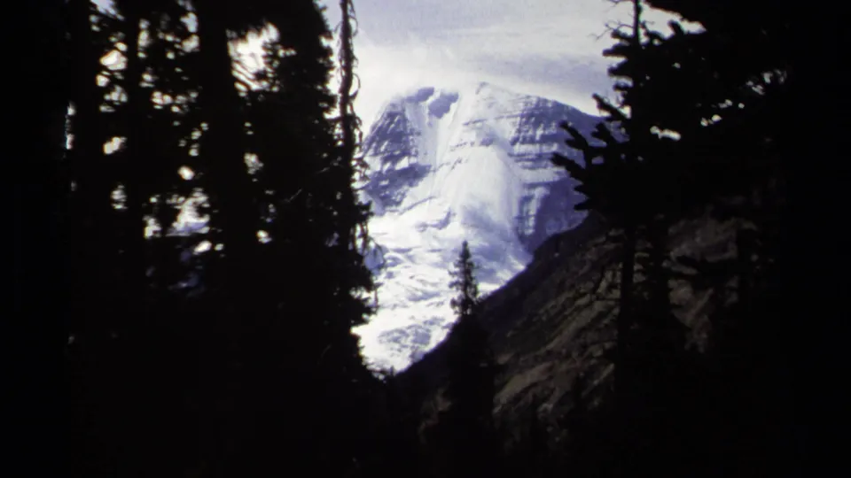 A glacier-covered mountain face appears framed by dark pines in British Columbia home-movie footage from 1979.