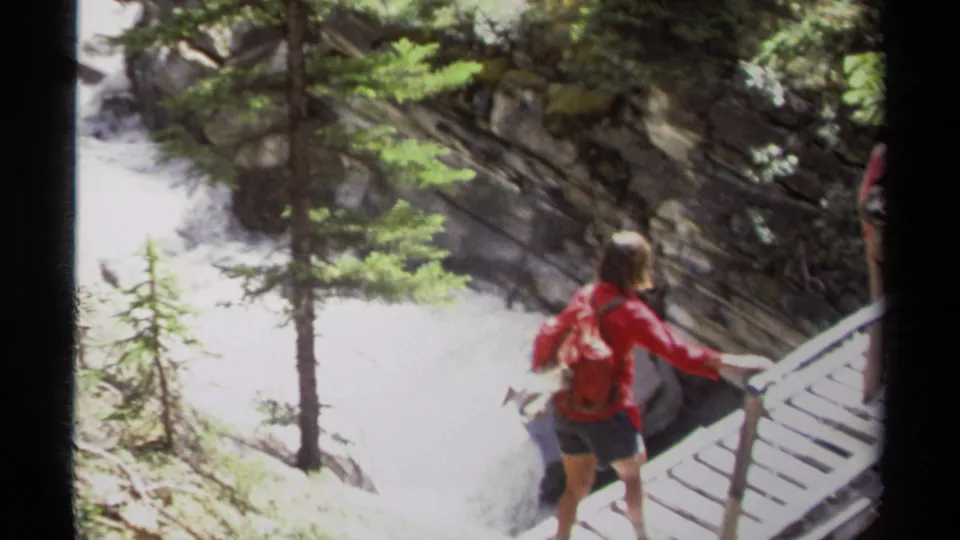 A distant hiker in red crosses a wooden boardwalk beside rushing water in 1979 British Columbia 8mm footage.