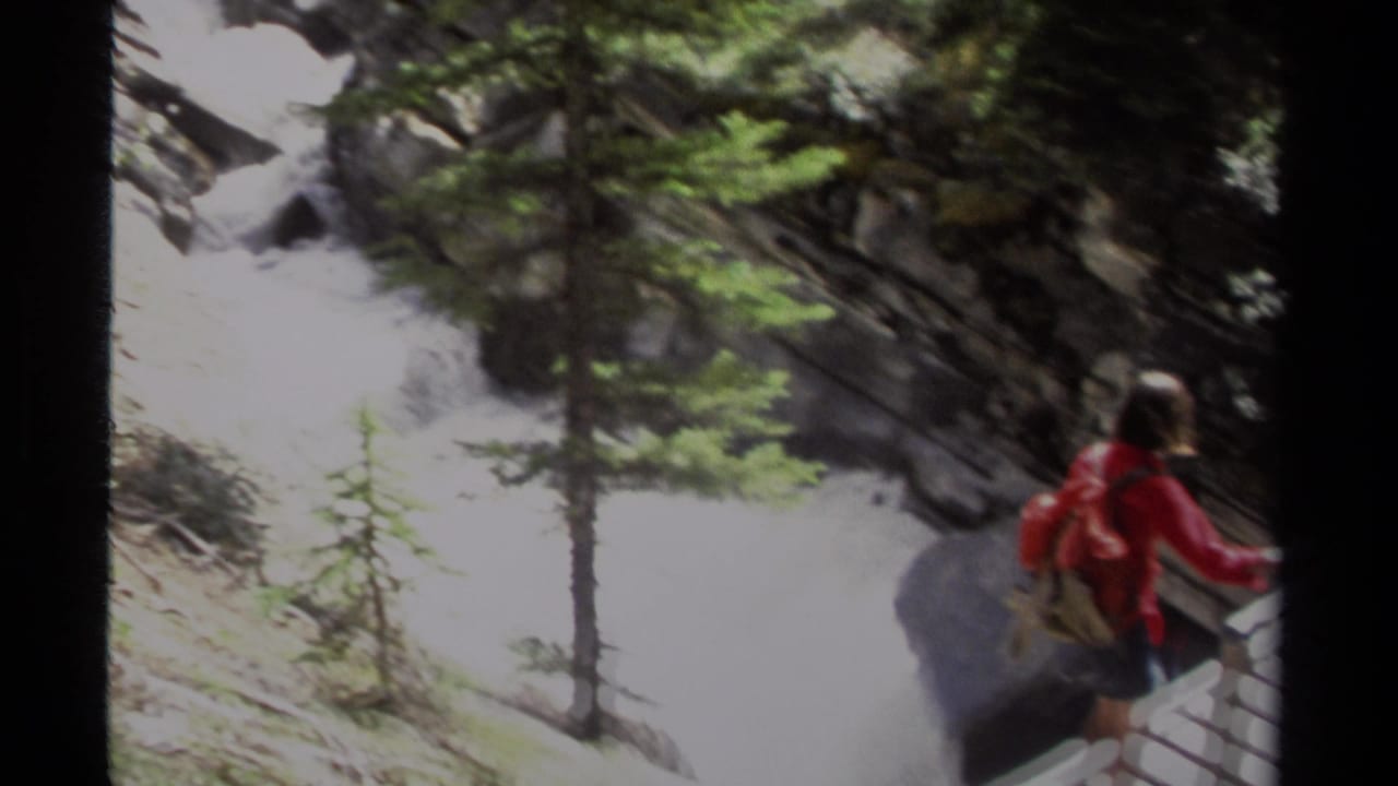 Woman in a red jacket crossing a wooden bridge above rushing water in a forest.
