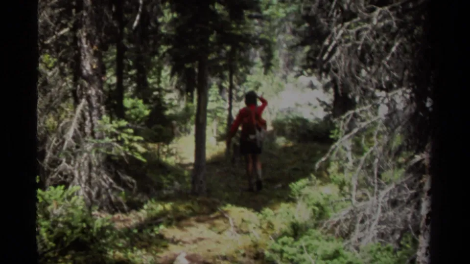 A woman in a red jacket and backpack walks away along a shaded British Columbia forest path in 1979 home-movie footage.