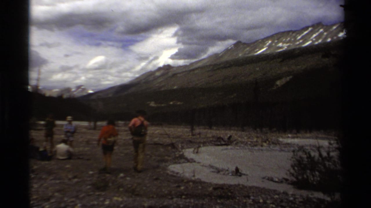 1979 British Columbia 8mm frame showing hikers in a rocky mountain valley near shallow water and distant snow patches.