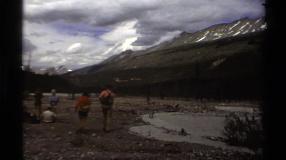 1979 British Columbia 8mm frame showing hikers in a rocky mountain valley near shallow water and distant snow patches.