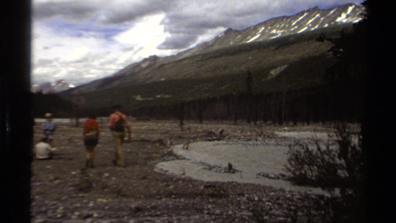 Hikers in British Columbia appear in a rocky alpine valley with a small pond, forest edge, and snow-streaked mountains.