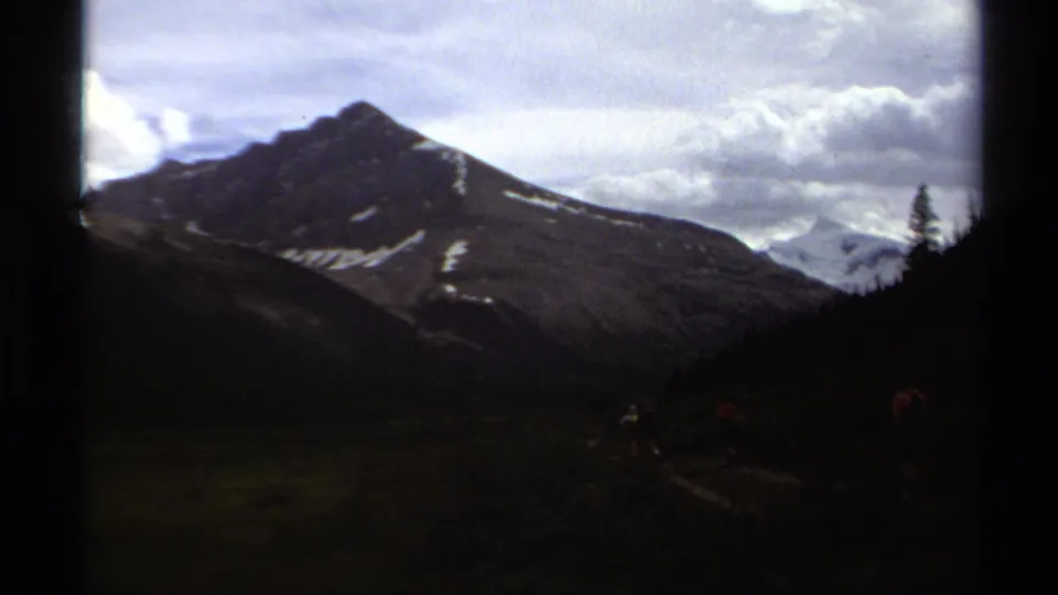 1979 British Columbia home-movie frame with hikers low in the scene below a dark mountain peak and cloud cover.