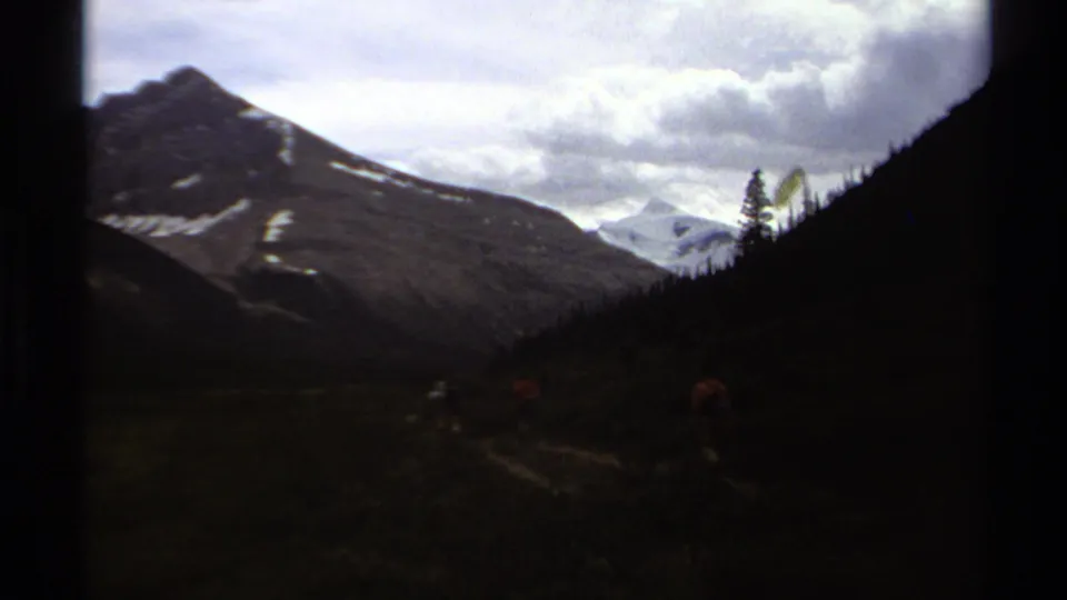 British Columbia 8mm footage frame showing hikers moving through a shadowed valley beneath snow-streaked mountains.