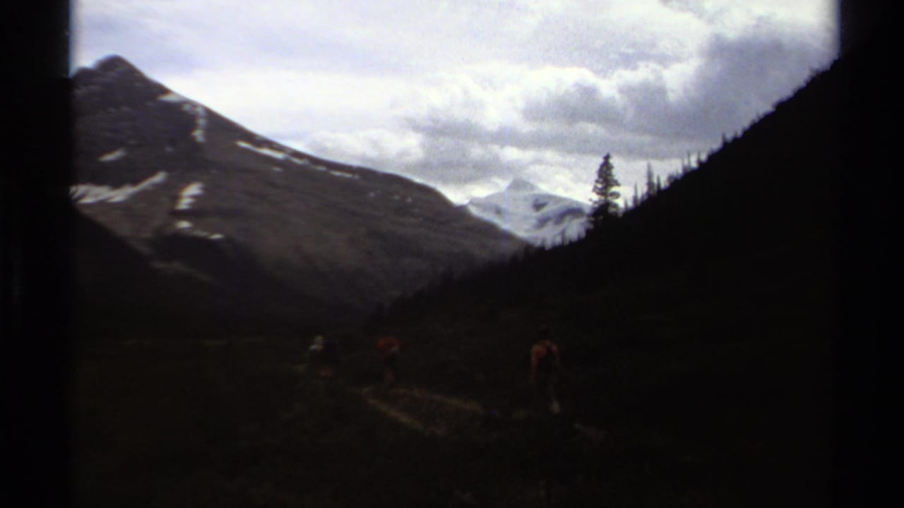 Hikers move through a dark mountain valley below cloudy sky and snow-capped peaks.