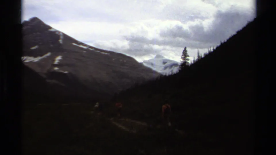 1979 8mm British Columbia footage showing distant hikers on a valley trail with forested slopes, clouds, and snow-capped mountains.