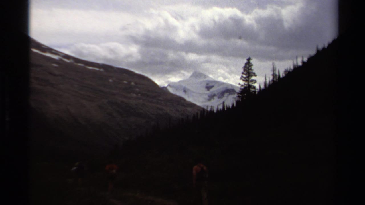 Small hikers walk in a dim valley framed by forested slopes and distant snowy mountains.