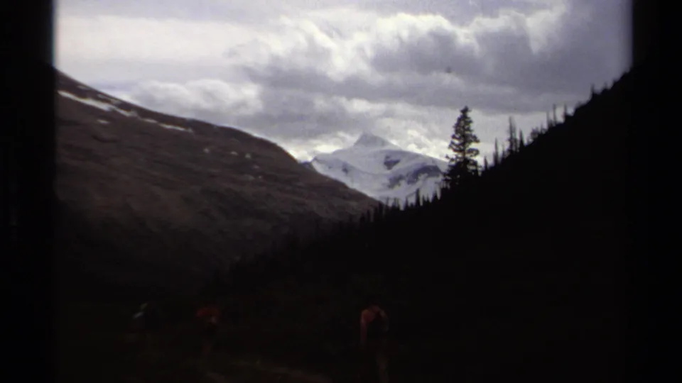 Distant hikers appear on a British Columbia mountain trail, with dark evergreen slopes and snow-covered peaks under heavy clouds.