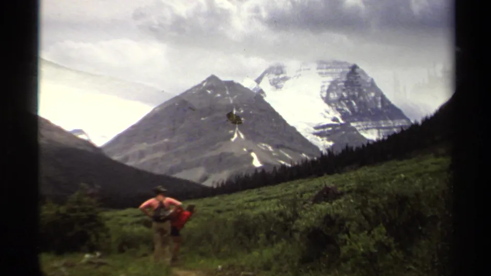1979 home-movie frame of hikers with backpacks in a green British Columbia meadow below snow-streaked alpine peaks.