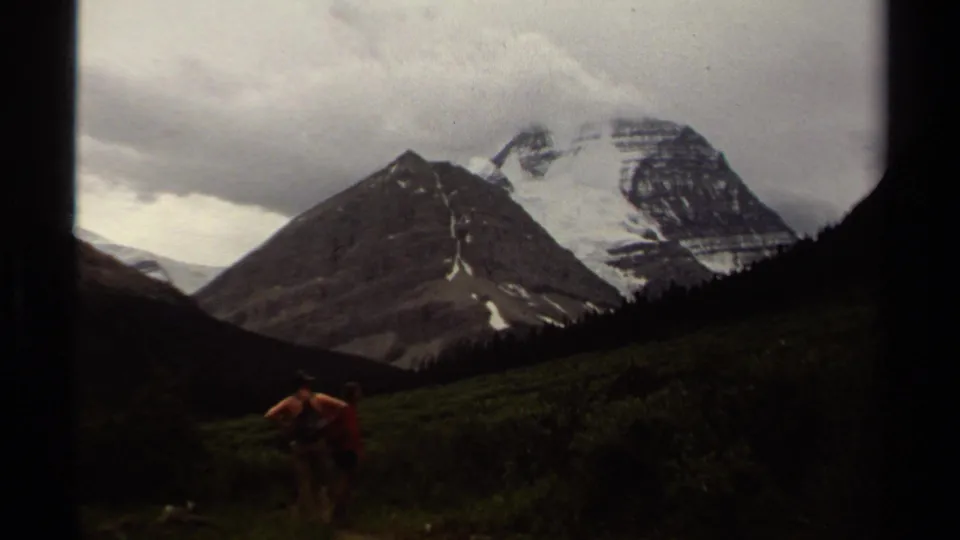 British Columbia 8mm footage shows hikers in an alpine meadow facing rugged mountain slopes and a clouded snow-capped summit.
