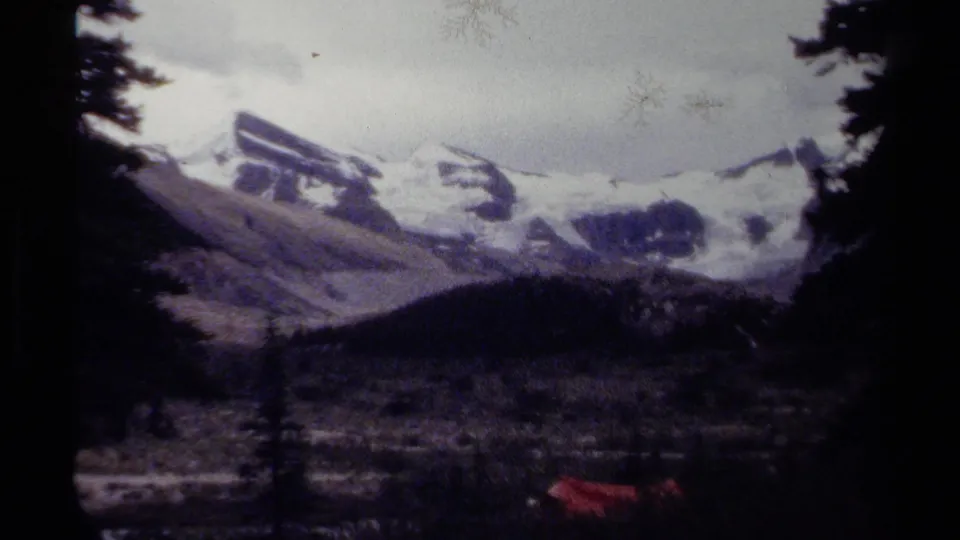 Soft 8mm frame of British Columbia mountains, showing snowy ridges, cloudy sky, dark trees, and a red object low in frame.