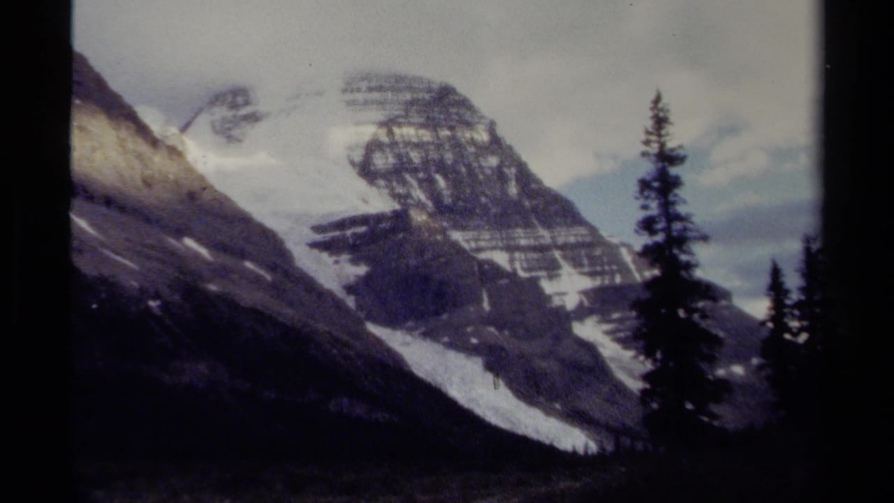 1979 8mm British Columbia mountain view with a snow-streaked rocky peak, dark conifers, and soft overcast light.